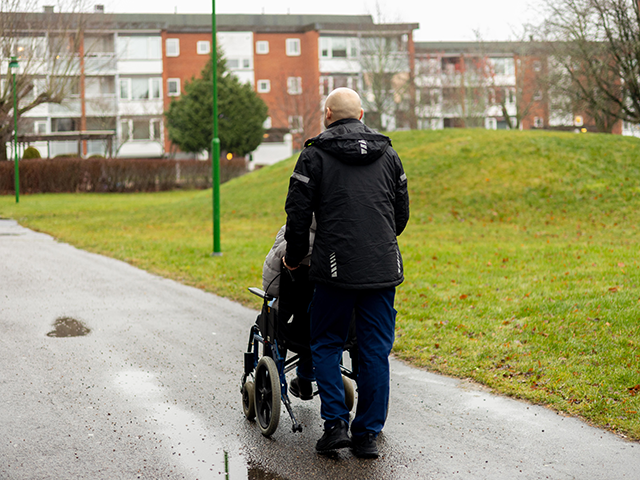 En person klädd i omsorgens arbetskläder skjutsar en person i en rullstol.