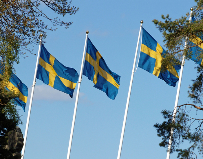 Ett fotografi på fem vajande svenska flaggor på flaggstångar, med blå himmel bakgrunden.