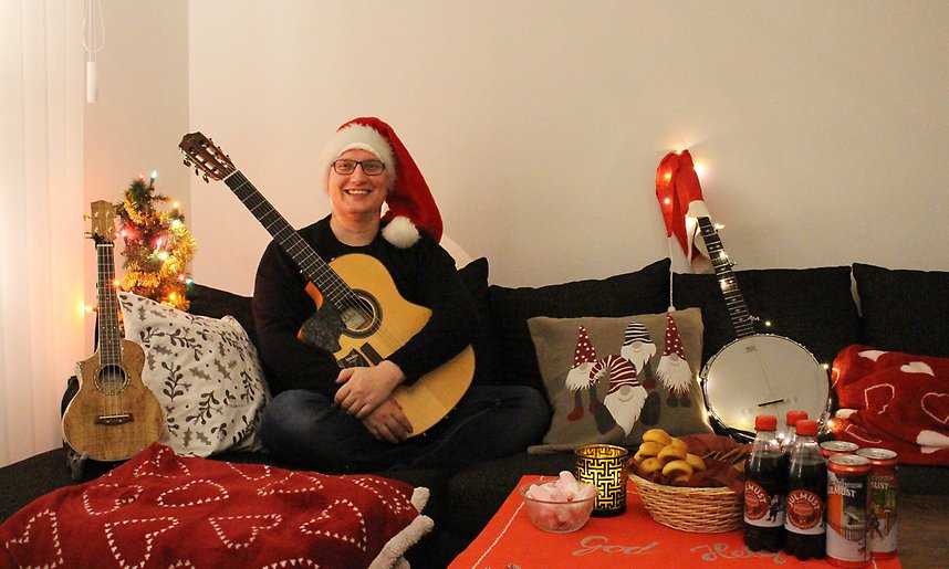En man sitter i en soffa med en gitarr i famnen och omgiven av juldekorationer.
