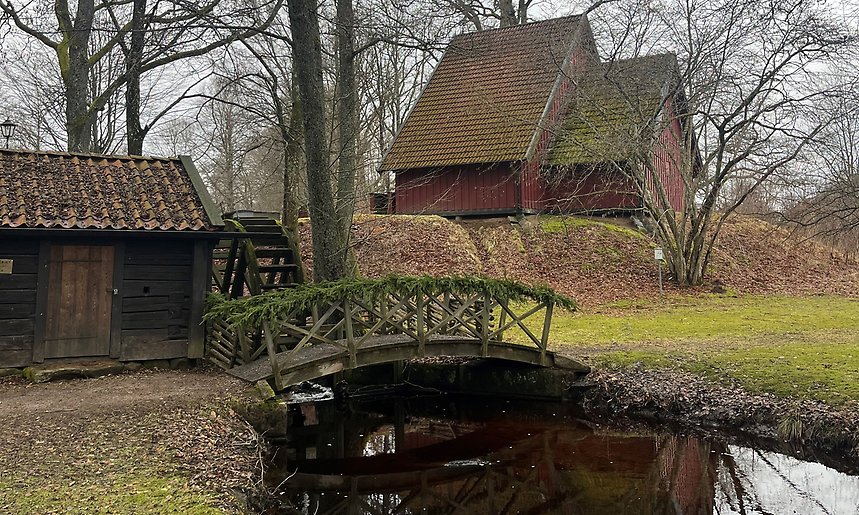 Bild på Hembygdspark där man ser två hus vid en sjö och en liten bro 