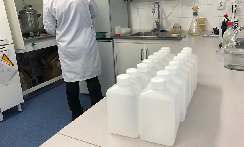 I förgrunden står plastflaskor på rad, i bakgrunden syns ryggen på en person i vit rock som jobbar i ett laboratorium.