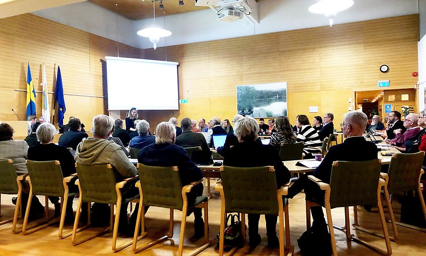 Kommunfullmäktiges ledamöter sitter i sina stolar, i bakgrunden syns en bildskärm