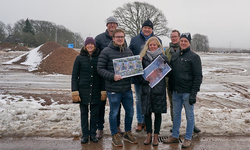 Sju personer står på två rader i grupp. Två av dem håller bilder med skisser på flerbostadshus i händerna. I bakgrunden syns ett markberett område och en stor ek.