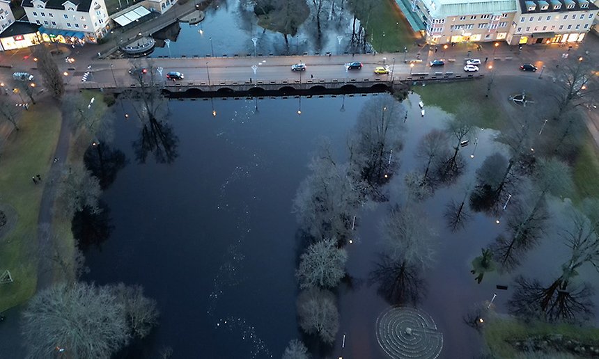 Bild från ovan som visar Åbron i mitten, där kör det bilar. Vattnet sår högt i ån. Det syns gröna områden i kanterna.