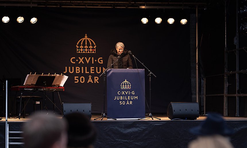En kvinna står bakom ett podium på en scen. På podiet och i bakgrunden syns en symbol som består av en kungakrona och texten C.XVI.G Jubileum 50 år.