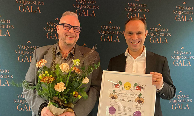 Två personer står intill varandra, en av dem håller en diplom, den andre håller en bukett blommor.