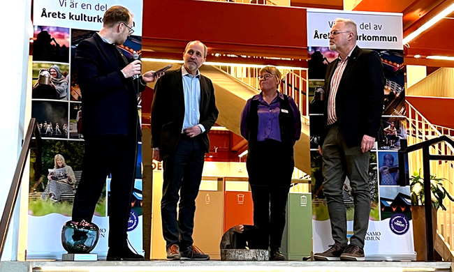 Fyra personer står på ett podium, en av dem håller i en mikrofon. Längst fram till vänster står ett äpple i glas.