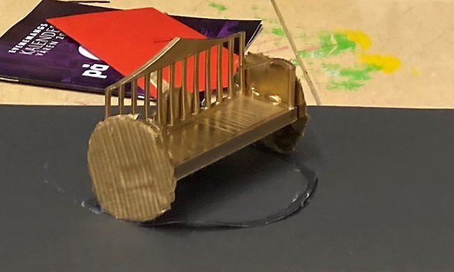 En minimodell av en bänk som ser ut som om flera pinnastolar ställs intill varandra och byggts ihop. Den är mässingsfärgad.