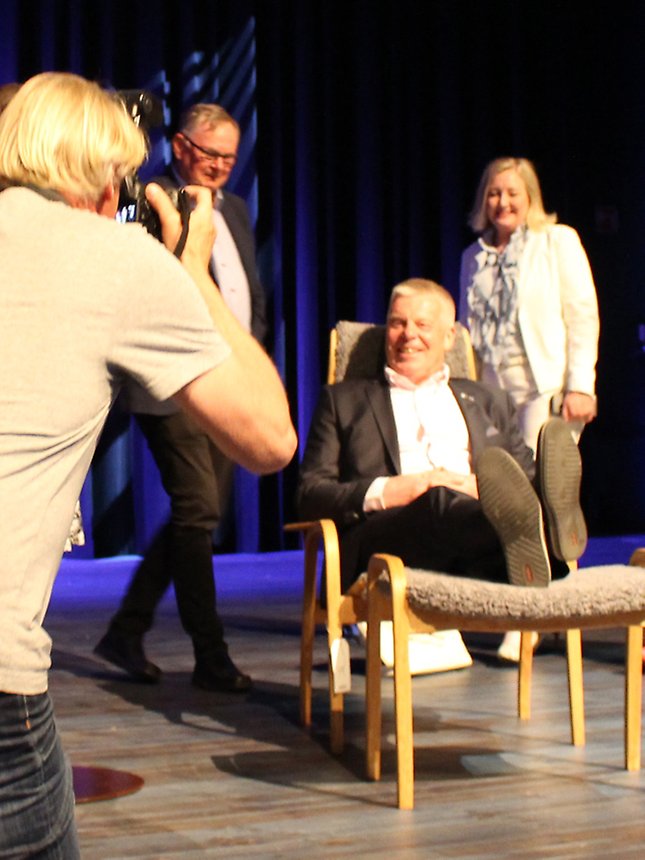 Bilden visar Hans-Göran Johansson, som sitter i en stol, som han fått av kommunstyrelseordförandena i länet. Värnamo Nyheters fotograf tar en bild.