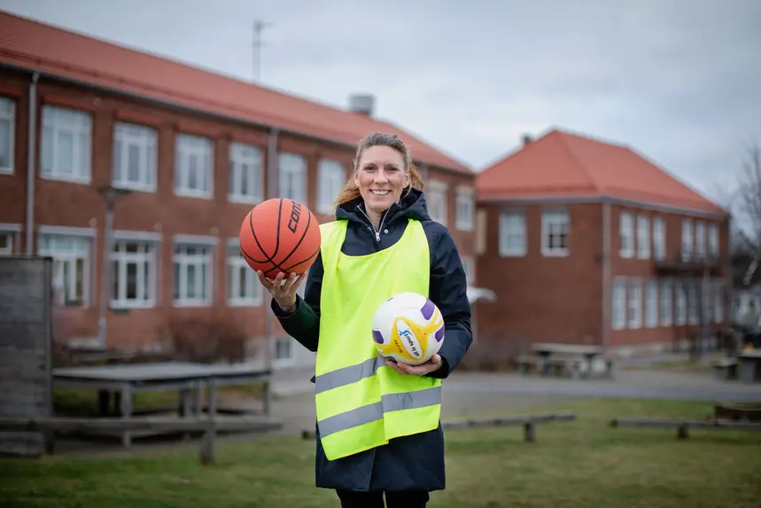 En kvinna klädd i ytterkläder och gul väst håller en boll i handen. I bakgrunden syns en skola.